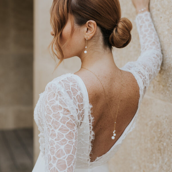 Mariée portant des bijoux de mariage élégants et minimalistes en perles d'eau douc et porcelaine. Elle porte un collier de dos monté sur une fine chaîne, et des boucles d'oreilles pendantes.