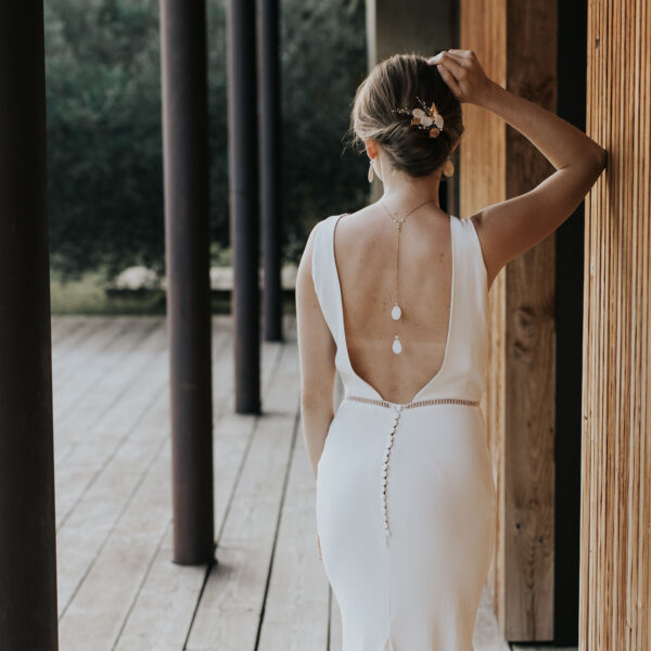 Mariée portant ses bijoux de mariage en porcelaine minimalistes et élégants. Elle porte des boucles d'oreilles et un pendentif de dos sur sa robe dos nu et des épingles à chignon dans sa coiffure de mariée.
