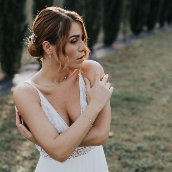 Portrait d'une mariée, la main posée sur l'épaule. Elle porte plusieurs bijoux de mariage dont un bracelet, un collier et des boucles d'oreilles en perles d'eau douce et perles bleu opal, montés sur une fines chaîne.